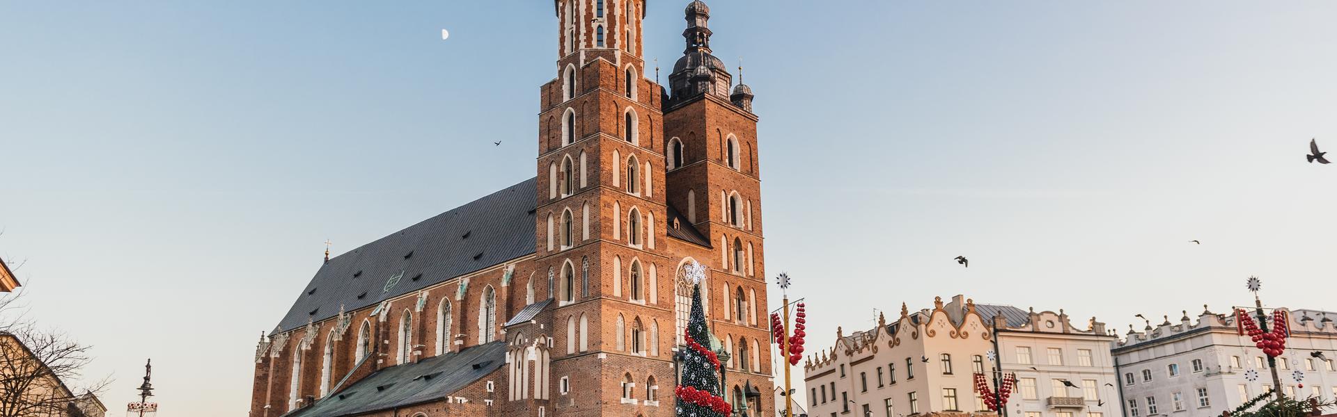 Przed kościołem mariackim na krakowskim rynku stoi wysoka choinka z czerwonymi i białymi ozdobami oraz drewniane stragany.