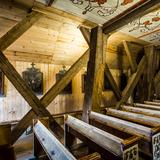 Wnętrze drewnianego kościoła - rząd drewnianych ławek zdobionych malowidłami, na drewnianych ścianach wiszą obrazy - stacje Drogi Krzyżowej, na suficie proste malowidła.