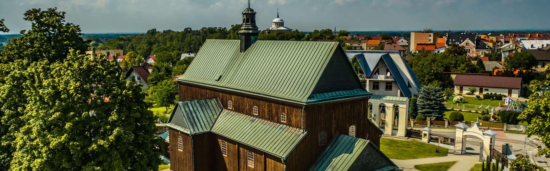 View of the wooden church in Dąbrowa Tarnowska. There are trees around the building. City buildings in the background.