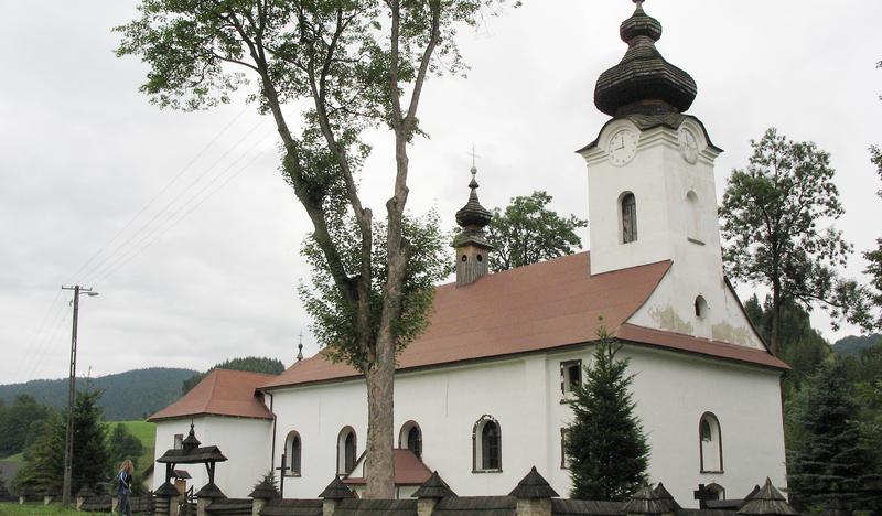 Kościół o prostej bryle z wieżą z cebulastym hełmem, biały z miedzianymi dachami. Otoczony kamienno-drewnianym ogrodzeniem.