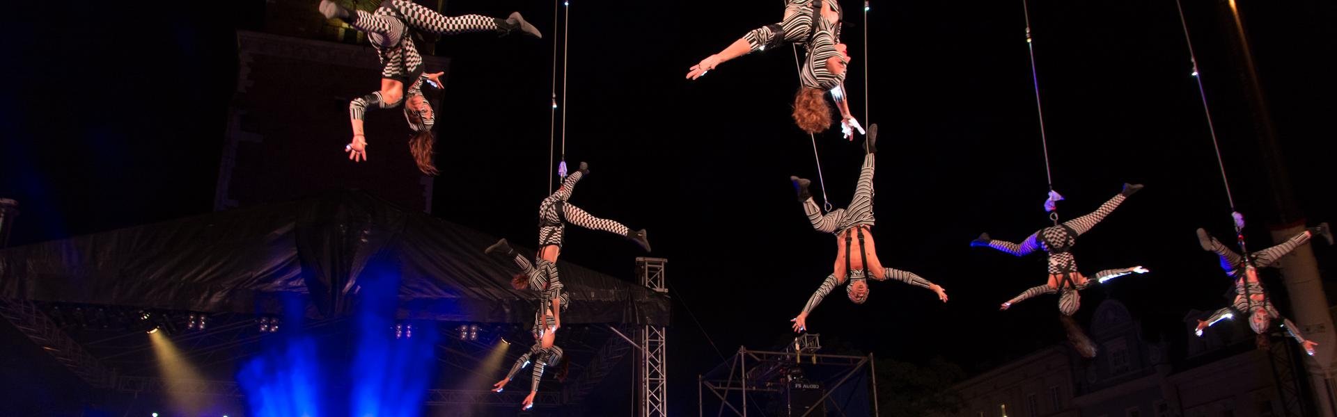 Disguised actors hanging upside down on ropes above the audience during a theater festival. It is dark, next to it there is a stage lit in blue.