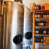 Zdjęcie przedstawia na pierwszym planie wysokie metalowe tanki przeznaczone na wino, zaś w tle półkę z licznymi produktami-butelkami zawierającymi wino które pochodzi z winnicy słońce i wiatr.