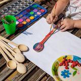 Ręce dziecka trzymają pędzelek i malują farbami drewnianą łyżkę, obok leżą farbki, inne łyżki i talerzyk z poziomkami