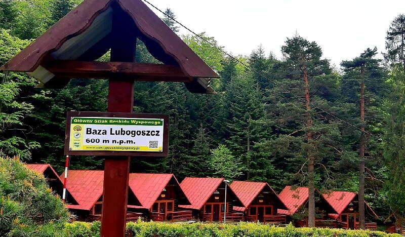 Drogowskaz Baza Lubogoszcz. Na drugim planie szereg drewnianych domków. W tle gęsty, zielony las.