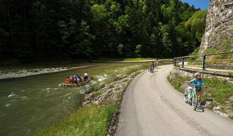 Zakręt asfaltowej drogi biegnącej pomiędzy wysoką skałą a rzeką Dunajec, na drodze rowerzyści prowadzący rowery, na rzece łódź flisacka z turystami