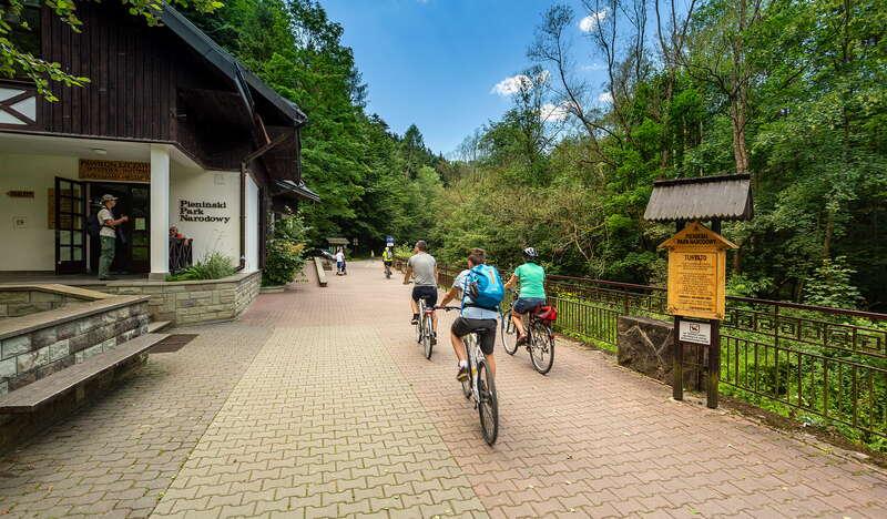 wybrukowana droga biegnąca obok pawilonu Pienińskiego Parku Narodowego, po której poruszają się rowerzyści, po prawej stronie drogi drewniana tablica z napisem 