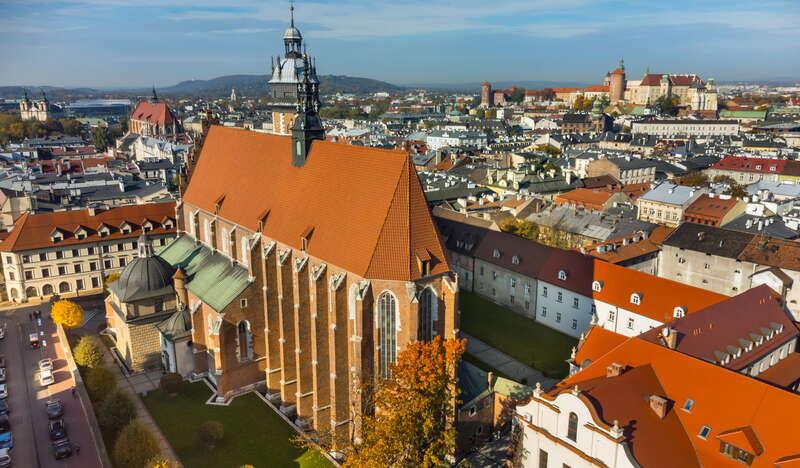 Widok na ceglaną, strzelistą Bazylikę Bożego Ciała. Wokół niej znajdują się budynki z ceglanymi dachami. W oddali jeszcze więcej budynków oraz Wawel.