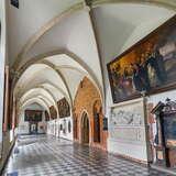 Korytarz w budynku Bazyliki Trójcy Świętej w Krakowie. Białe ściany zdobią duże obrazy, a sufity zdobią żebrowe sklepienia.